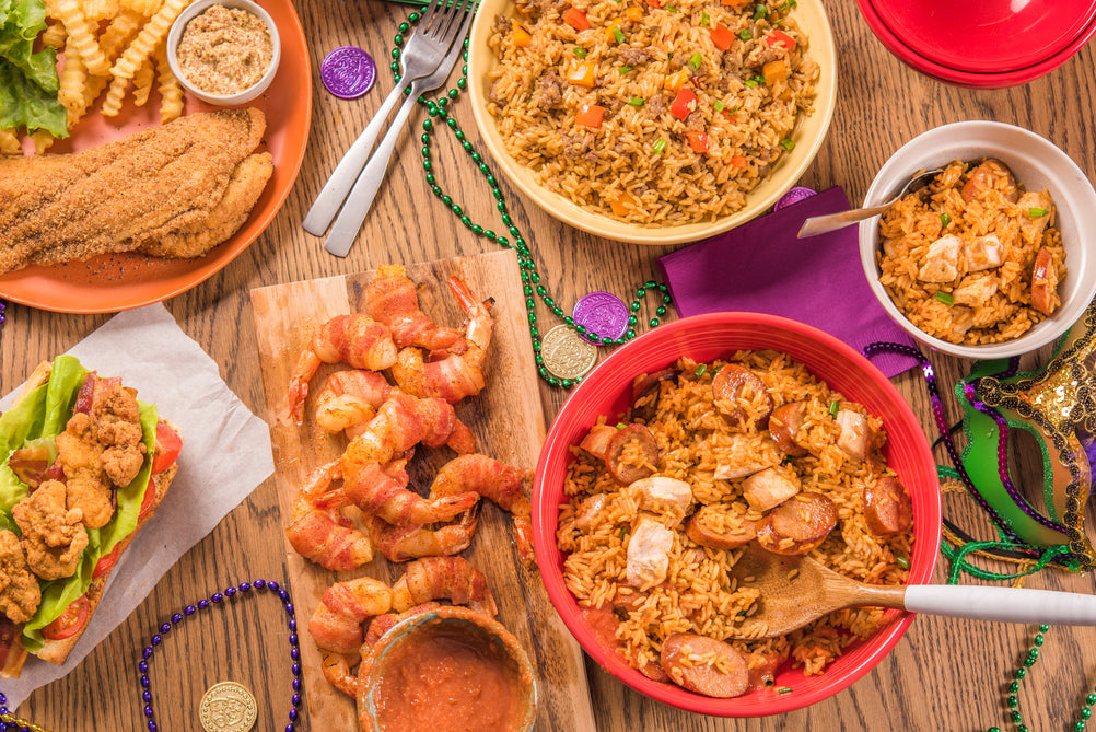 Diverse food items including fried shrimp, jambalaya, and other dishes on a wooden table with Mardi Gras decorations.